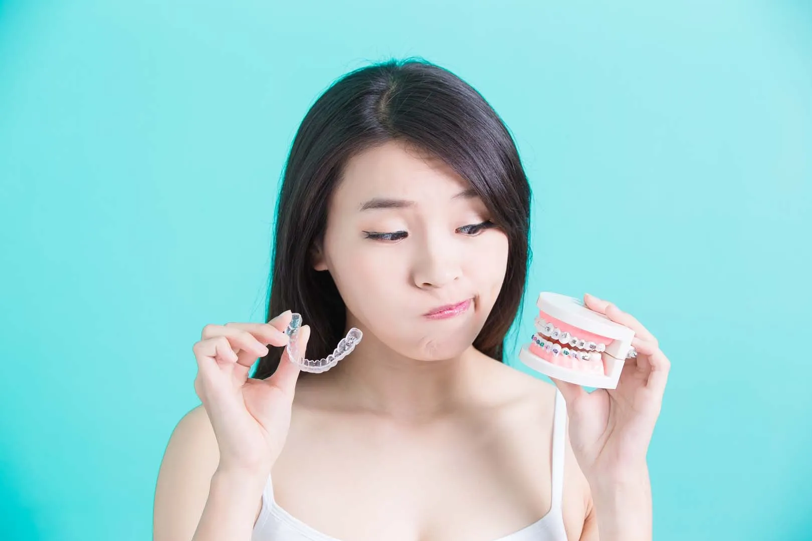 Woman holding clear dental aligner in one hand and dental braces model in the other, looking unsure.