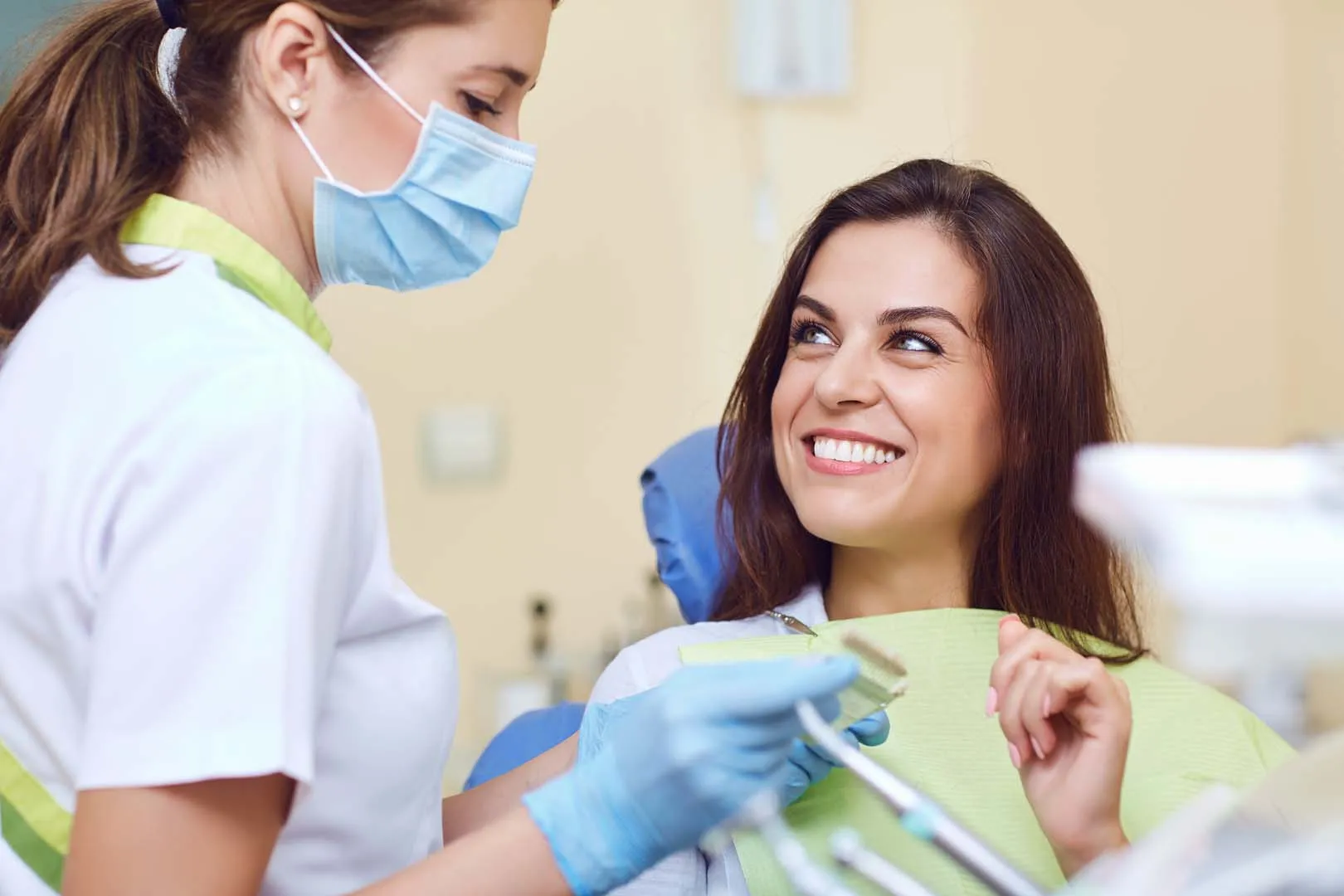 Female dentist wearing mask and gloves talks to smiling woman patient in dental chair.